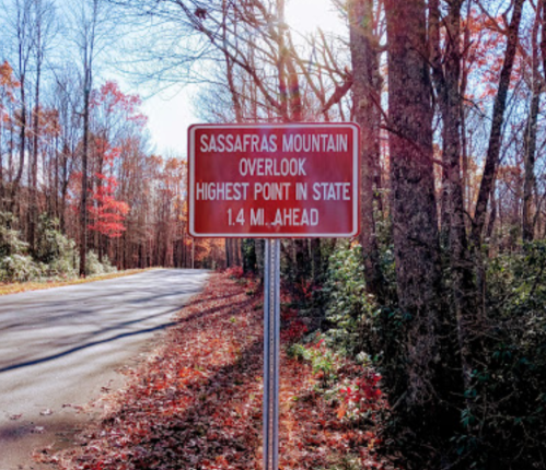 Sign for Sassafras Mountain Overlook, indicating it's the highest point in the state, 1.4 miles ahead. Autumn foliage visible.