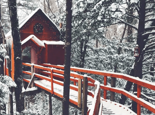 A cozy red treehouse surrounded by snow-covered trees, connected by a wooden walkway.