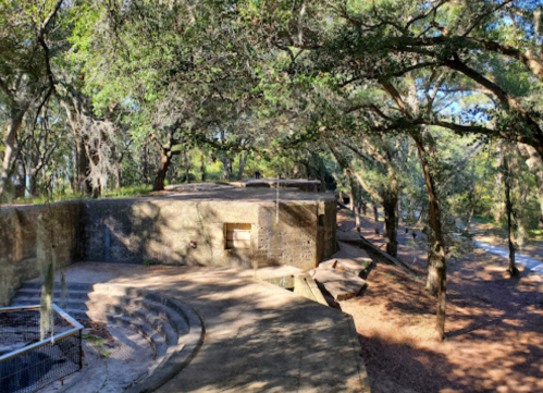 A stone structure surrounded by trees, featuring steps and a pathway in a wooded area. Sunlight filters through the leaves.