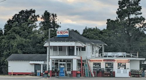 A vintage Exxon gas station with a two-story building and a convenience store, surrounded by trees under a cloudy sky.