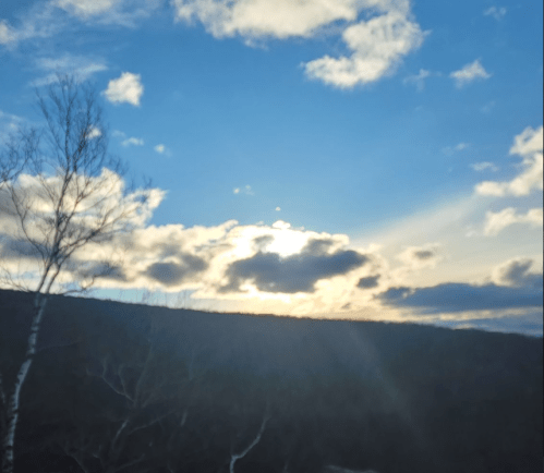 A serene landscape featuring a bright sky with clouds and the sun setting behind a distant mountain.
