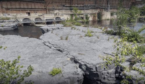 Rocky terrain with cracks, surrounded by water and sparse vegetation, near an industrial building.