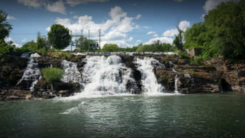 A serene waterfall cascading over rocky cliffs into a calm river, surrounded by lush greenery and a blue sky.