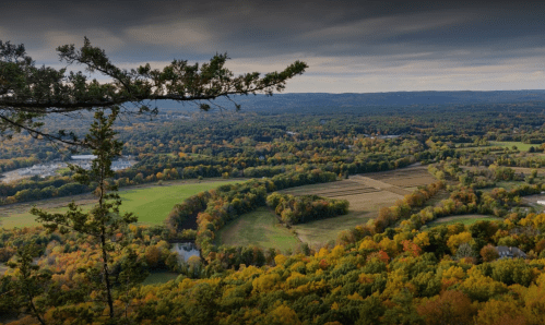 A scenic view of rolling hills and a river, surrounded by vibrant autumn foliage under a cloudy sky.