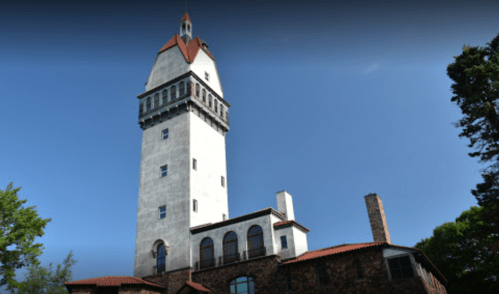A tall stone tower with a red roof, surrounded by trees and a blue sky.
