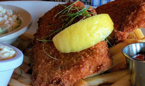 A plate of crispy fried fish with a lemon wedge, served with fries and a side of coleslaw and dipping sauce.