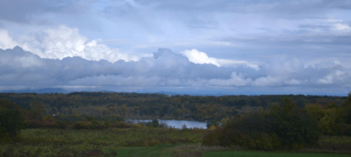 A scenic view of a river surrounded by trees and rolling hills under a cloudy sky.
