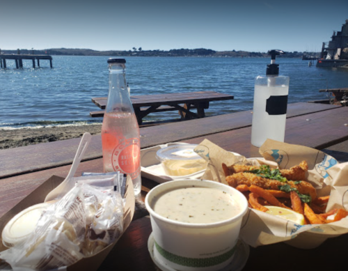 A scenic view of a waterfront meal featuring fries, clam chowder, and a drink on a sunny day.