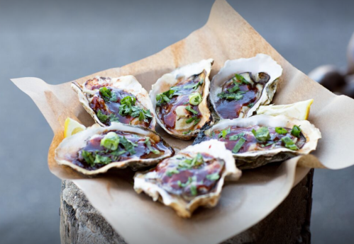 A plate of six grilled oysters topped with green onions and sauce, served on a rustic wooden surface.