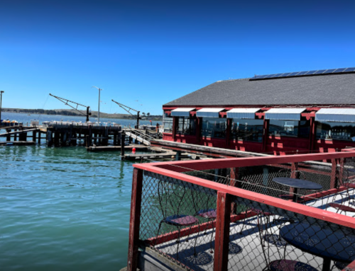A waterfront restaurant with red accents, overlooking a calm bay and fishing docks under a clear blue sky.