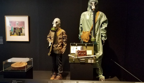 A child-sized mannequin in a gas mask and jacket stands next to an adult mannequin in a protective suit, with artifacts displayed.