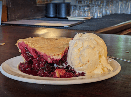A slice of berry pie with a golden crust served alongside a scoop of vanilla ice cream on a white plate.