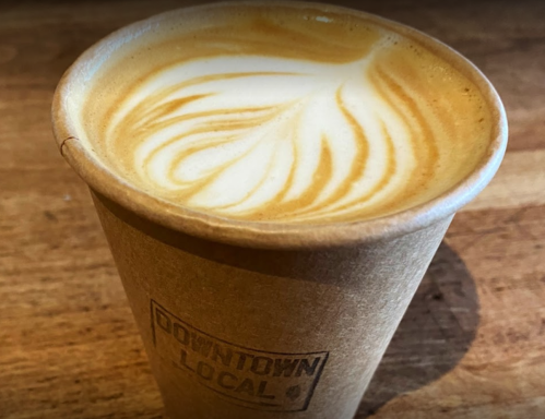 A latte in a brown paper cup with intricate foam art on top, resting on a wooden surface.