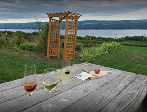 Three glasses of wine on a wooden table, with a scenic view of a lake and a wooden trellis in the background.