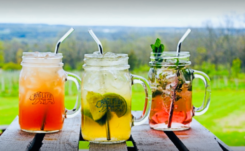 Three mason jars filled with colorful drinks, set on a wooden table with a scenic green landscape in the background.