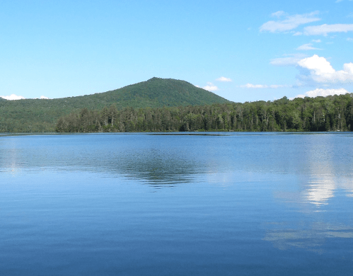 A serene lake reflects a green mountain under a clear blue sky with scattered clouds.