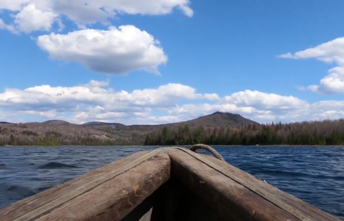 View from a boat on a lake, with blue skies, fluffy clouds, and distant mountains surrounded by trees.