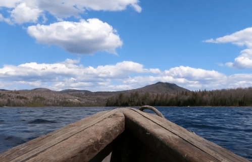 View from a boat on a lake, with blue skies, fluffy clouds, and distant mountains surrounded by trees.