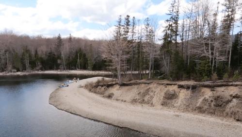 A serene riverbank scene with sandy shore, trees, and a few people enjoying the outdoors on a cloudy day.