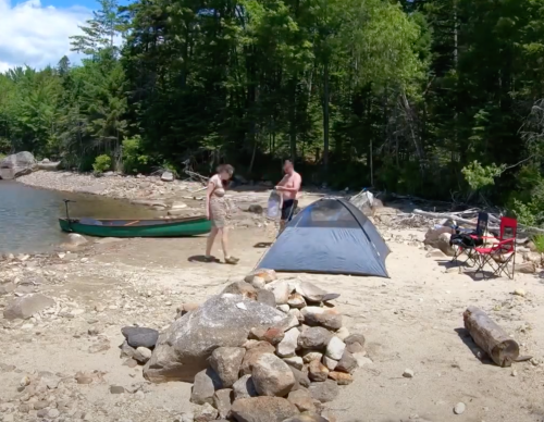 Two people set up a tent by a rocky lakeshore, with a canoe nearby and trees in the background.