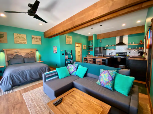 Cozy living space with a bed, sofa, and kitchen, featuring teal walls and wooden beams. Bright cushions add color.