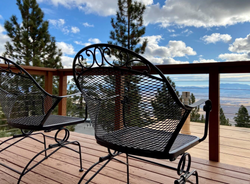 Two black metal chairs on a wooden deck, overlooking a scenic view of trees and mountains under a cloudy sky.