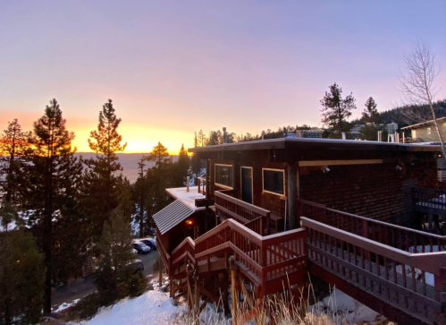 A cozy cabin at sunset, surrounded by snow and pine trees, with a view of a lake in the distance.