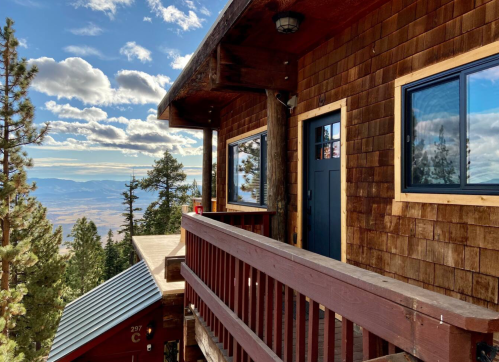 A wooden cabin with a balcony overlooks a scenic mountain view under a partly cloudy sky.