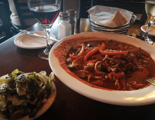 A plate of colorful stir-fried vegetables and meat in sauce, with a side of greens and glasses of red and white wine.