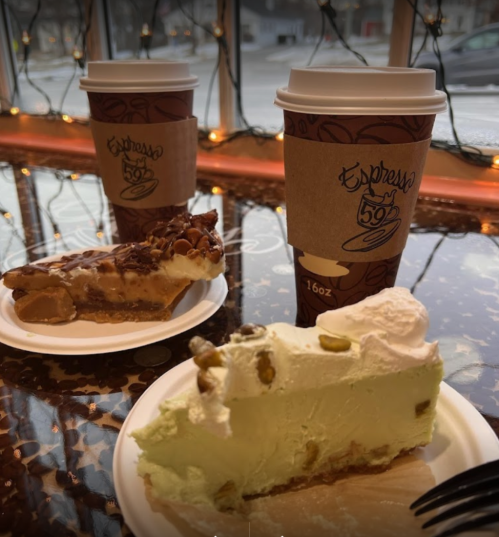 Two coffee cups and slices of pie on plates, with a cozy café atmosphere and festive lights in the background.