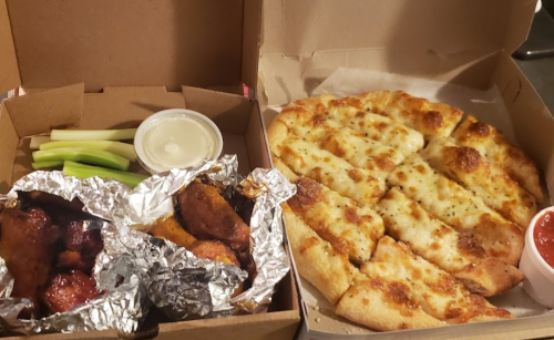 A takeout box with buffalo wings, celery sticks, and a side of ranch dressing next to a pizza with cheese and marinara sauce.