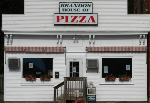 A white building with a sign reading "Brandon House of Pizza," featuring large windows and flower boxes.