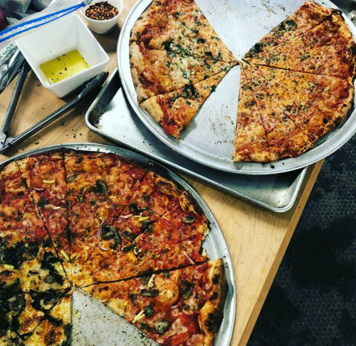 Two large pizzas on metal trays, one with various toppings and the other with cheese, alongside olive oil and spices.