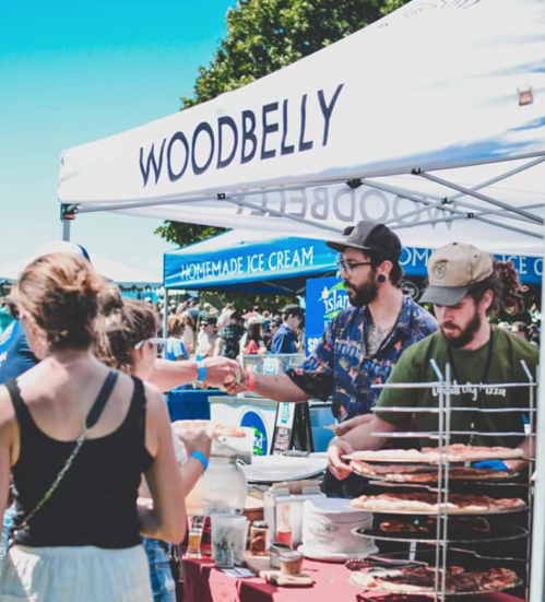 A busy outdoor market scene with people interacting at a food stall under a "Woodbelly" tent.
