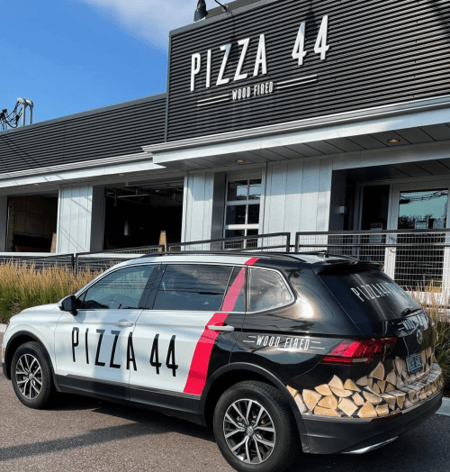 A car with "Pizza 44" branding parked outside a modern restaurant featuring a wood-fired pizza sign.