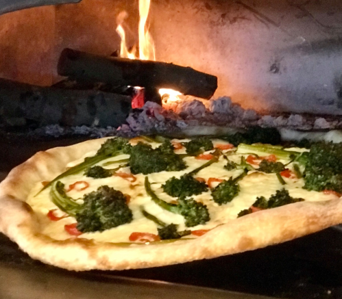 A freshly baked pizza with broccoli and green peppers, placed in a wood-fired oven with flames in the background.