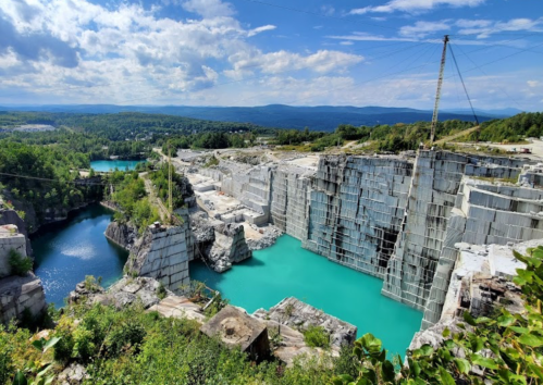 A scenic view of a quarry with turquoise water, surrounded by rocky cliffs and lush greenery under a blue sky.
