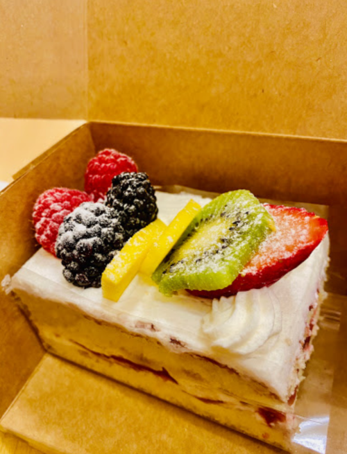 A slice of cake topped with raspberries, blackberries, kiwi, and lemon, in a brown takeout box.