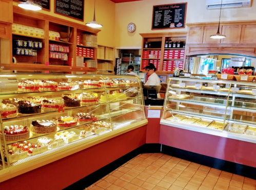 A bakery display filled with various cakes and pastries, with a staff member in the background.