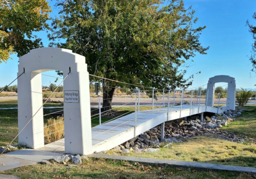 A small pedestrian bridge with white arches spans a rocky area, surrounded by grass and trees under a clear blue sky.