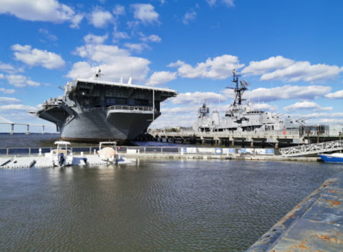 A large aircraft carrier docked alongside a smaller naval vessel under a blue sky with scattered clouds.