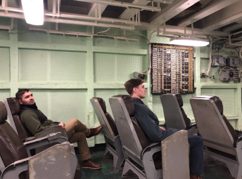 Two men sit in vintage chairs inside a green, industrial room with control panels on the wall.