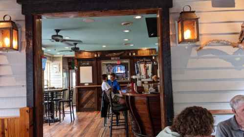 A cozy restaurant interior with wooden floors, a bar, and patrons seated at tables. Warm lighting and a relaxed atmosphere.
