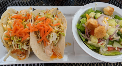 Two tacos filled with shredded lettuce, carrots, and fried shrimp, served with a side salad of mixed greens and croutons.