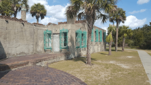 Ruins of a building with turquoise window frames, surrounded by palm trees and a paved walkway under a blue sky.