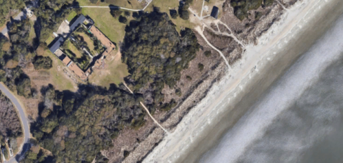 Aerial view of a coastal area with a sandy beach, greenery, and a building near the shoreline.
