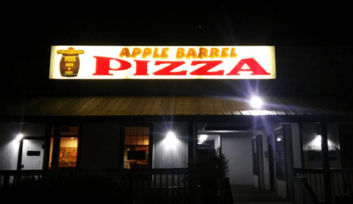 Sign for "Apple Barrel Pizza" illuminated at night, with a rustic building in the background.