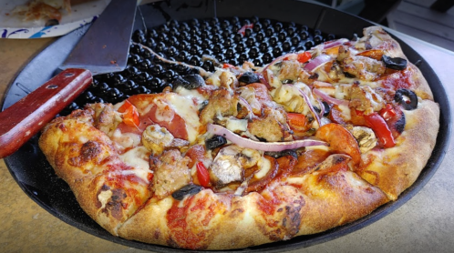A close-up of a pizza with various toppings, including sausage, peppers, and olives, on a black pizza pan.