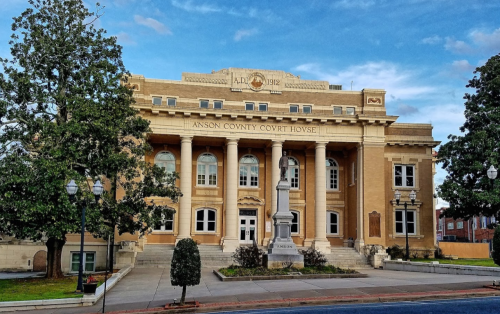 Historic Anson County Courthouse with a statue in front, surrounded by trees and a clear blue sky.