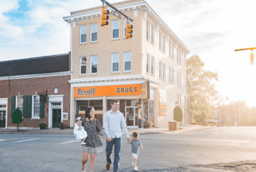 A family walks hand-in-hand near a drugstore on a sunny day, with a charming building in the background.